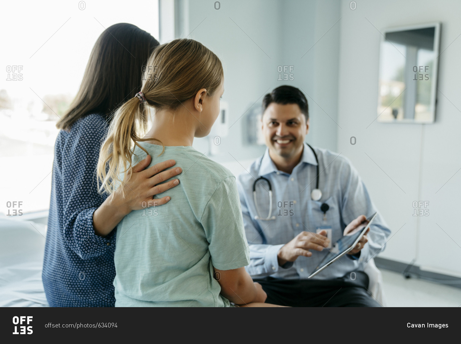 Cheerful pediatrician showing x-ray image on tablet computer to mother and daughter in medical examination room