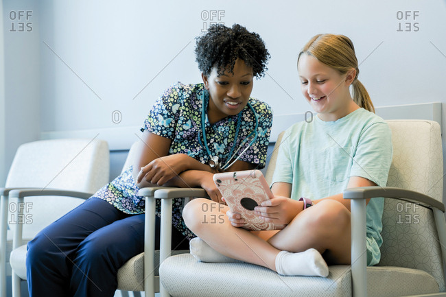 Pediatrician looking at girl using tablet computer while sitting on chair in hospital