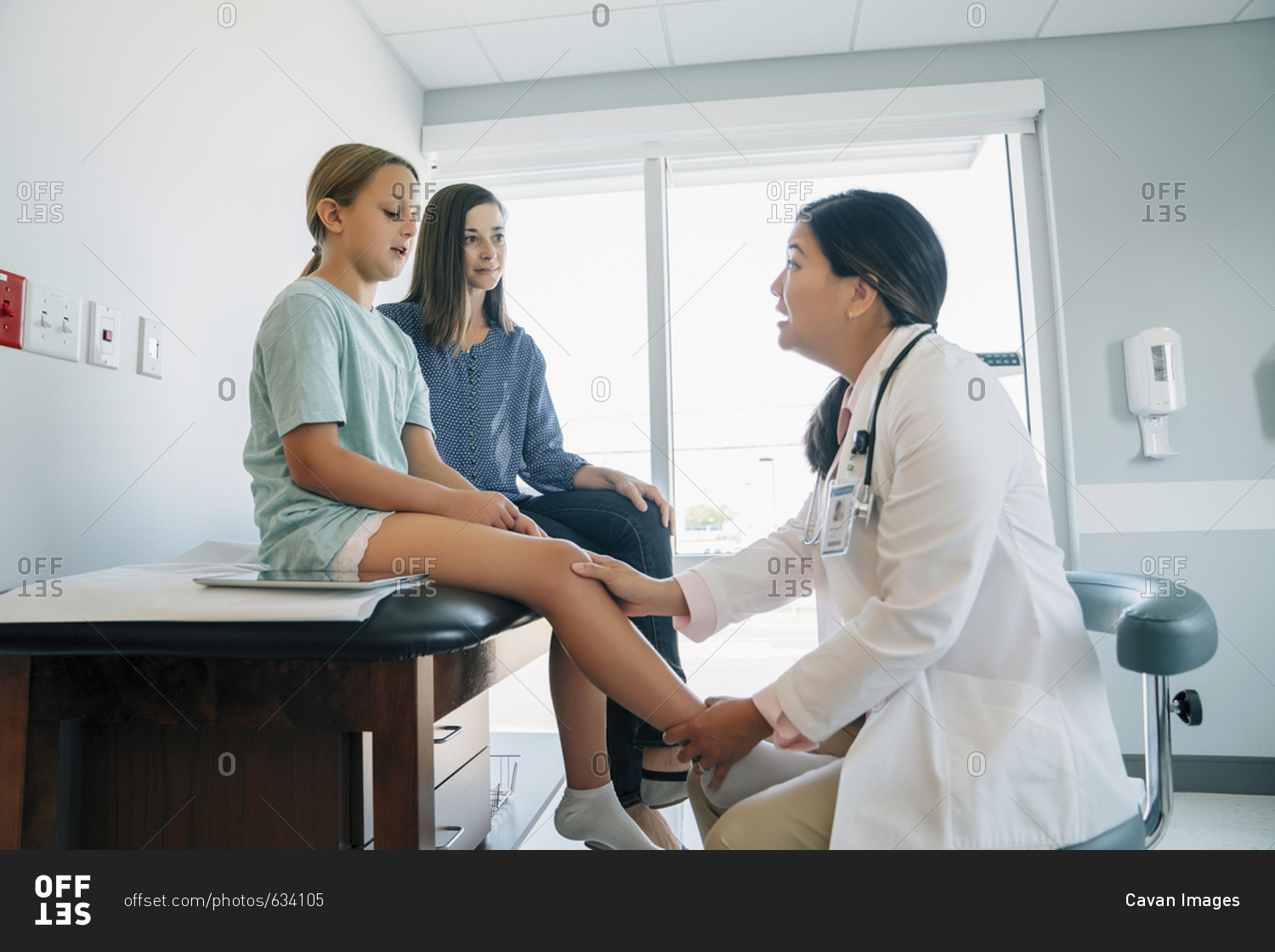 Mother looking at pediatrician checking daughter's knee joint in medical examination room