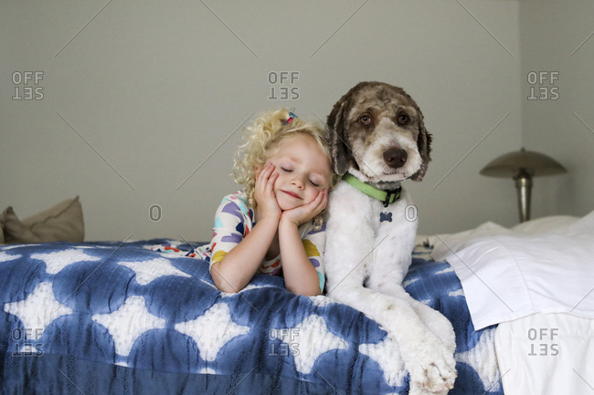 Girl with eyes closed and hands on chin lying by dog on bed against wall