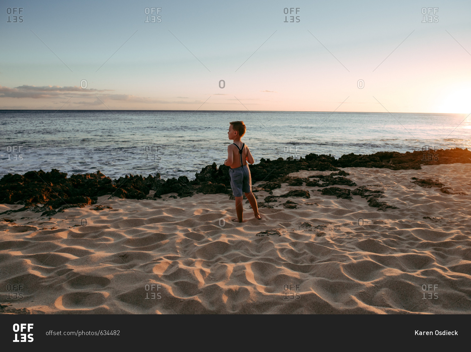 Boy wearing suspenders standing by the ocean at sunset
