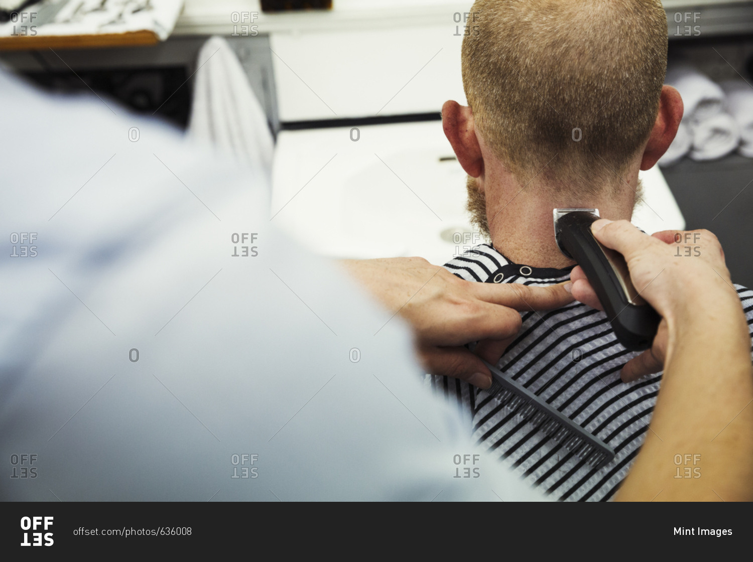 A customer sitting in the barber\'s chair, and a barber using a beard trimmer on his neck.