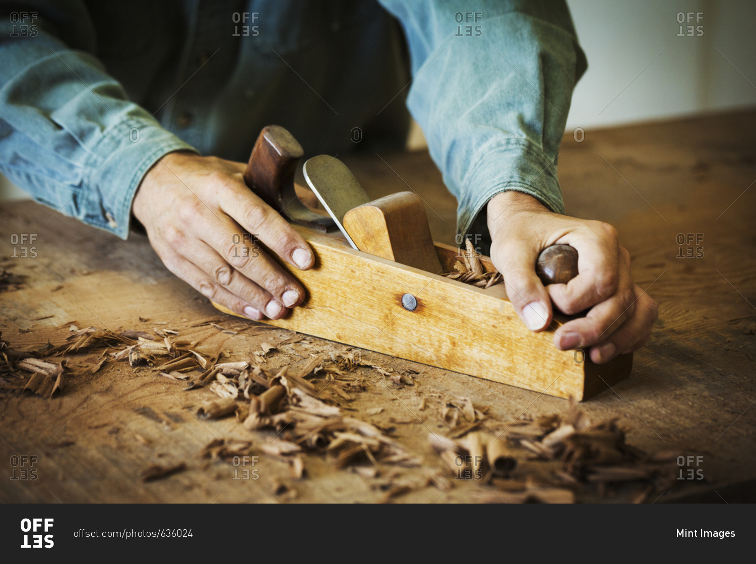 A workman using a hand wood plane on the surface of a large piece of wood.