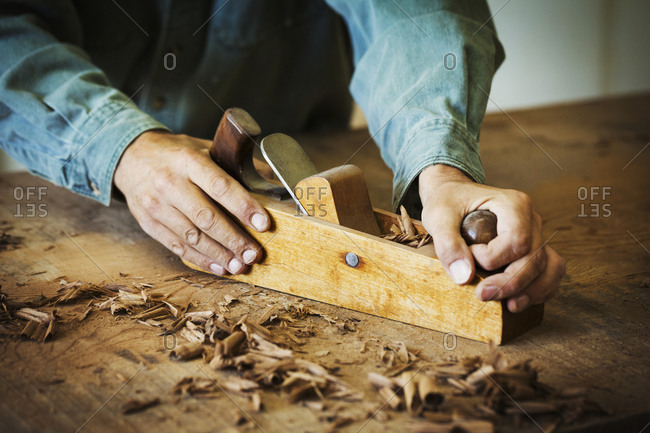 A workman using a hand wood plane on the surface of a large piece of wood.