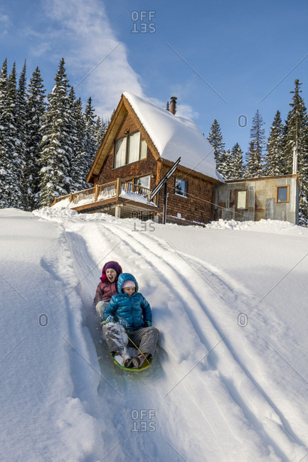 Two Young Girls Sledding In Powder Snow Below Mountain Cabin Near