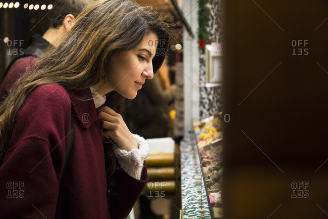 Beautiful young woman in red coat standing near stall at marketplace and choosing goods for buying.
