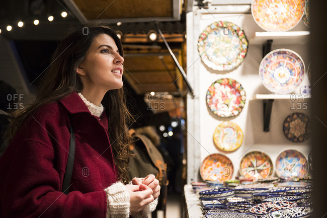 Beautiful woman in red coat standing at marketplace and looking at colorful patterned plates.