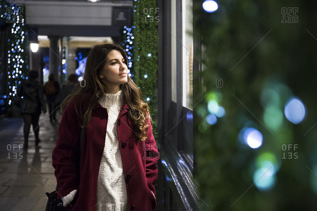 Charming woman in red coat walking near decorated shop and looking at shop-window.