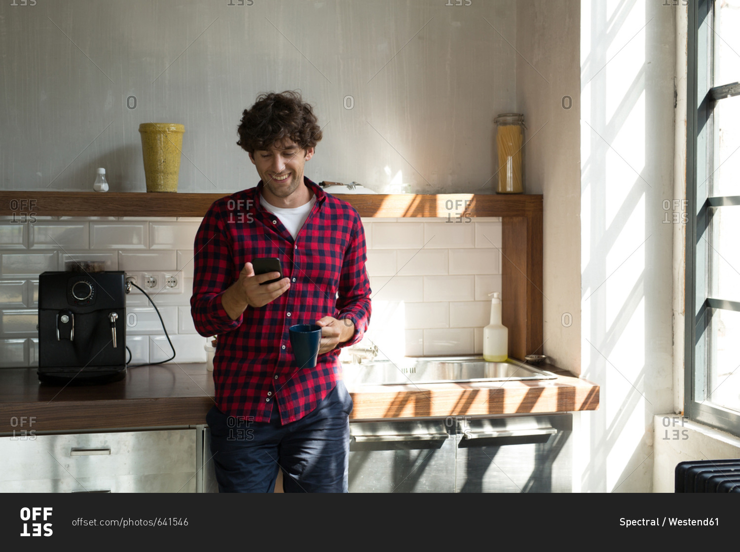 Young entrepreneur standing in company kitchen- drinking coffee- using smartphone