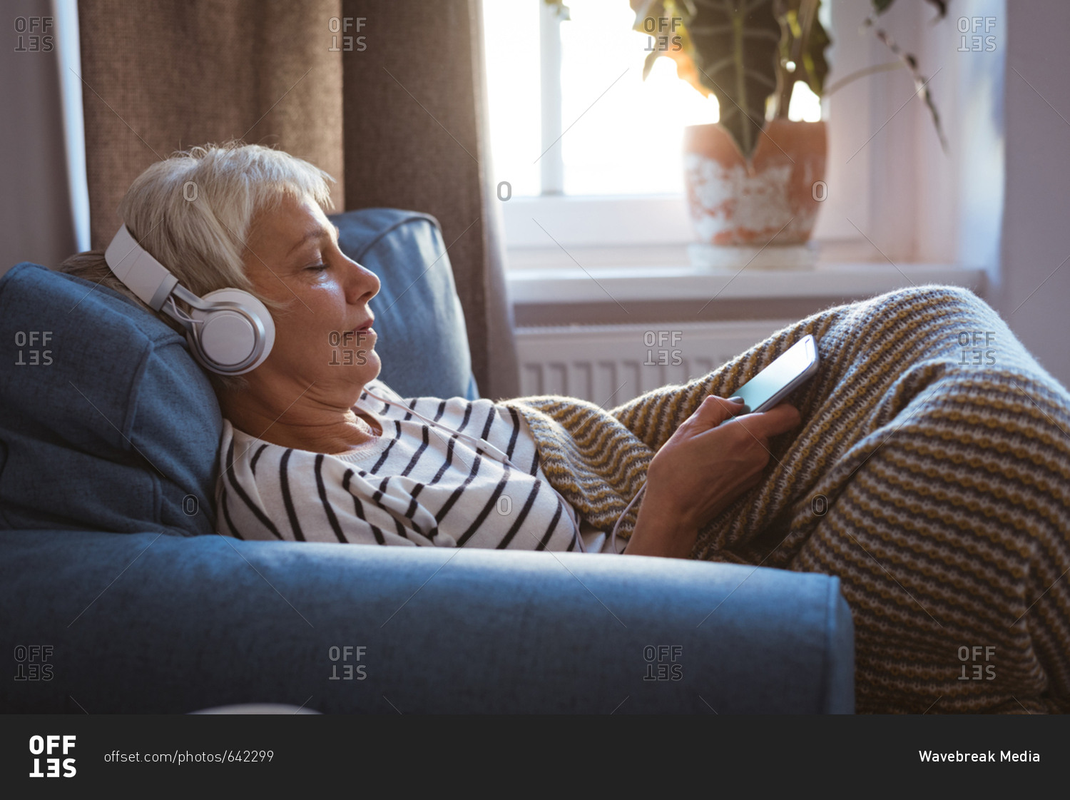 Senior woman listing to music on phone while siting on sofa in living room at home