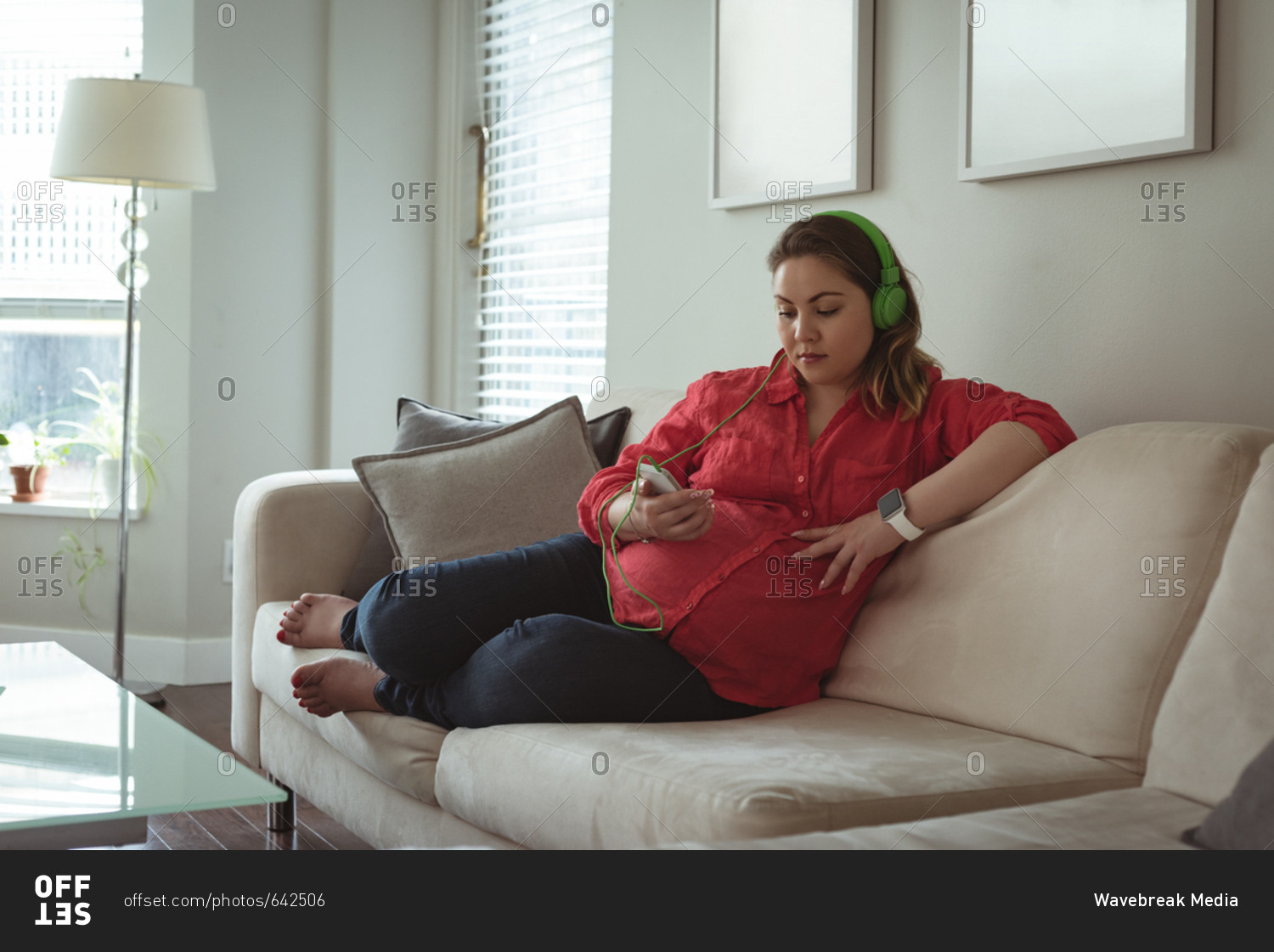 Young pregnant woman sitting on sofa listing to music on her mobile phone at home