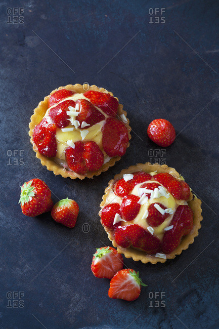 Two strawberry tartlets with custard and white chocolate shaving on dark ground