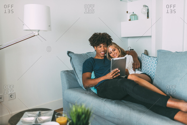 Happy young interracial couple relaxed at home lying down on the sofa looking at the tablet