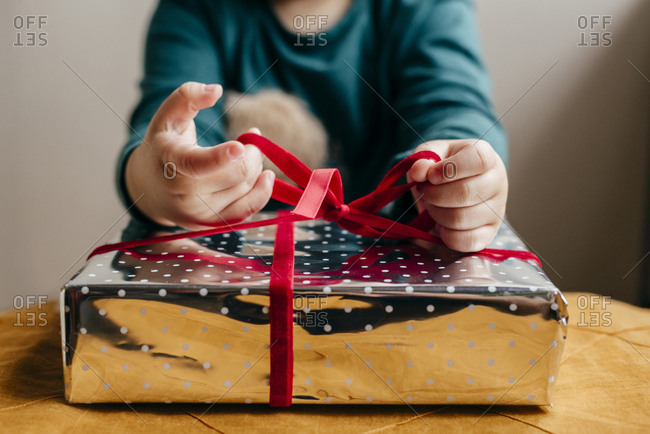 Crop child sitting at table and tying red ribbon on gift box wrapped in shiny paper