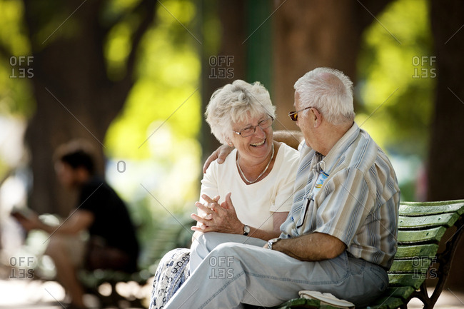 Senior woman smiles at a senior man with his arm around her shoulders as they sit together on a park bench