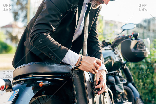 Mature businessman outdoors, leaning over motorcycle, doing up bag, mid section