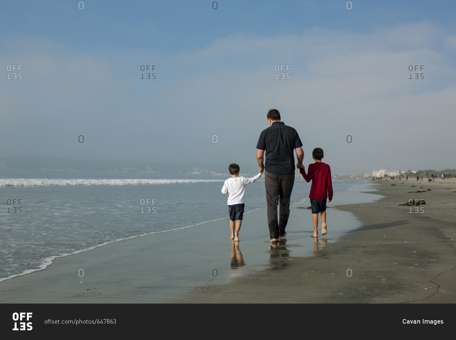 Rear view of father holding sons hands while walking on shore against sky at beach