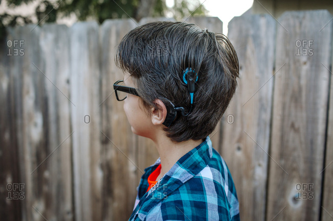 Side view of boy with hearing air standing against wooden fence at yard