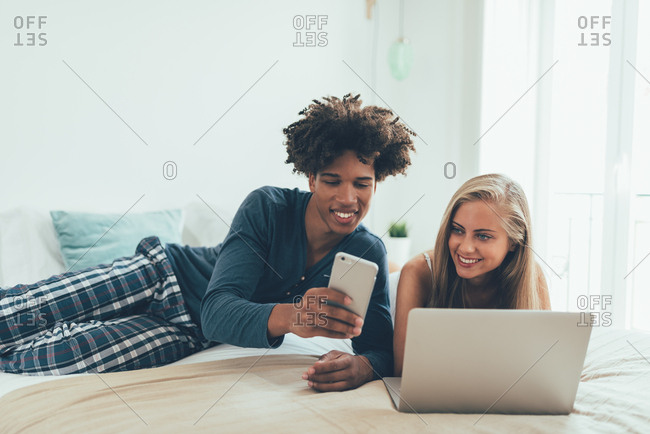 Young beautiful interracial couple relaxed lying down in bed on the computer and mobile phone