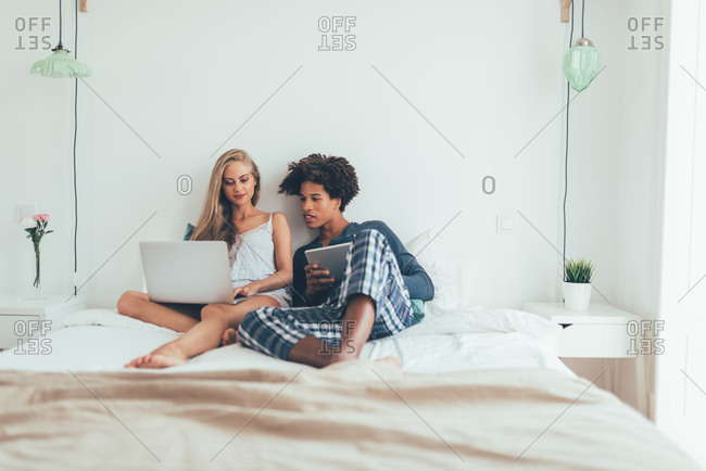 Young beautiful interracial couple relaxed lying down in bed on the computer and tablet