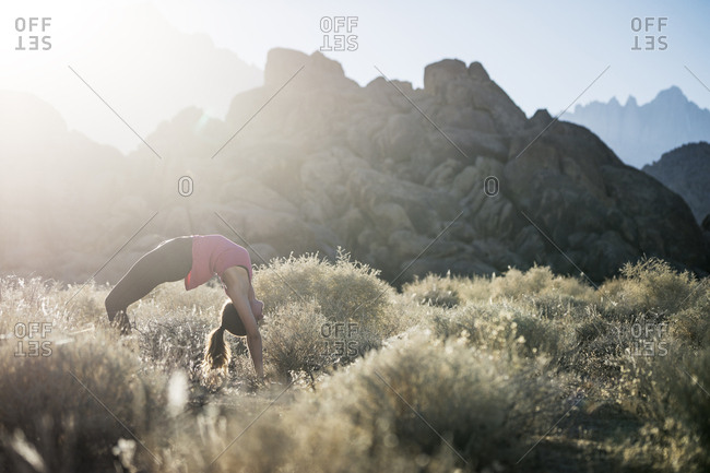 Woman bending over backwards while practicing yoga on field against mountains during sunny day