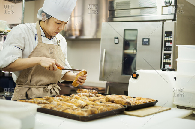 Woman wearing chef's hat and apron working in a bakery, slicing freshly baked rolls on large trays