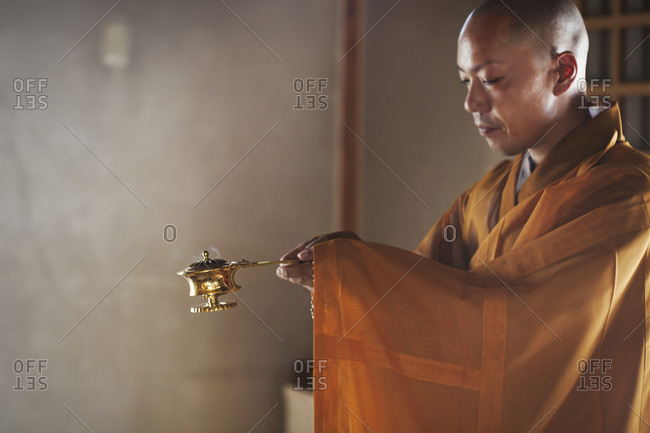 Side view of Buddhist monk with shaved head wearing golden robe kneeling indoors in a temple, holding incense burner