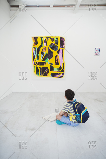 Boy with short black hair wearing backpack sitting on floor in art gallery with pen and paper, looking at modern painting