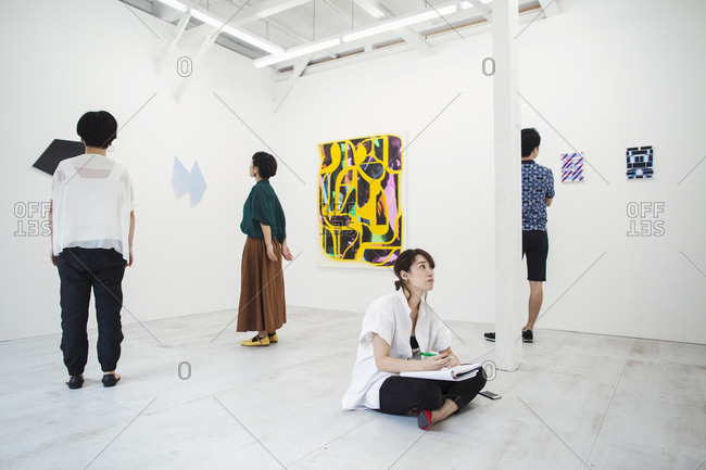 Woman with black hair sitting on floor in art gallery with pen and paper, looking at modern painting, three people standing in front of artworks