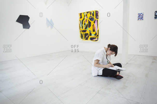 Woman with black hair wearing white shirt sitting on floor in art gallery with pen and paper