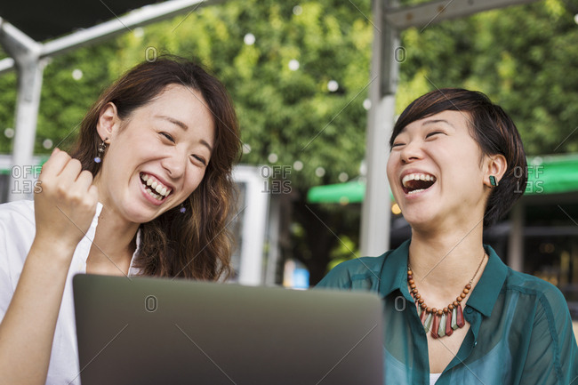 Two women with black hair wearing green and white shirt sitting in front of laptop at table in a street cafe, laughing