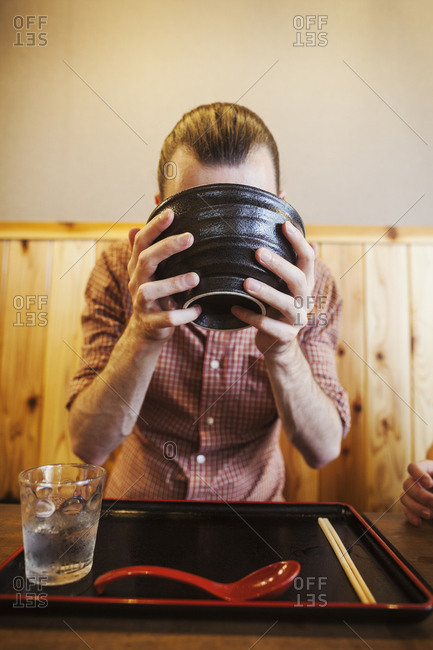 A western man in a noodle restaurant, with a noodle bowl lifting it with two hands to drink from it