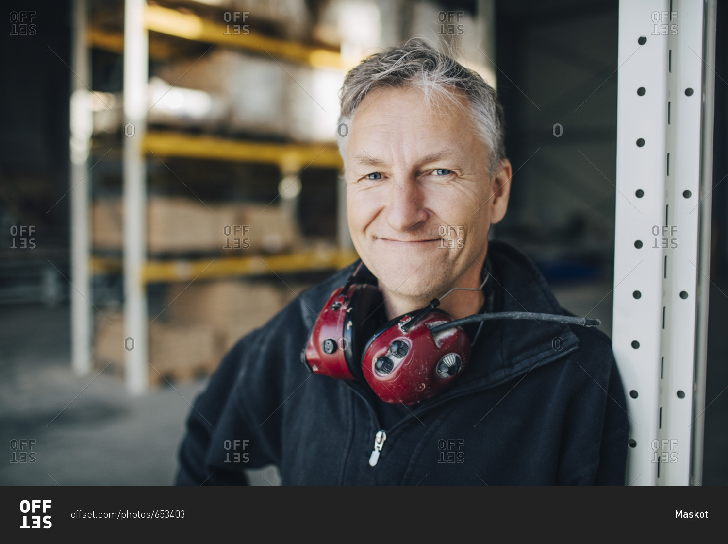 Close-up portrait of mature worker with ear protectors leaning on rack at industry
