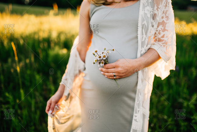 Expectant mother walking through sunlit field holding flowers and touching belly