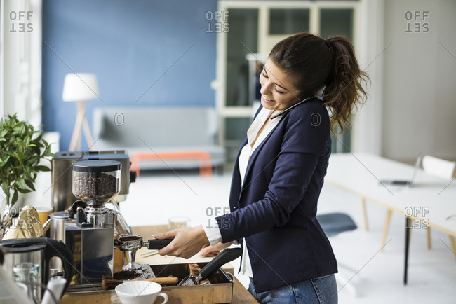 Smiling businesswoman on the phone preparing espresso with espresso machine in a loft