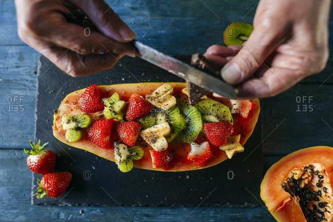 Close-up of man's hands cutting chocolate over half papaya garnished with pieces of banana- kiwi and strawberries