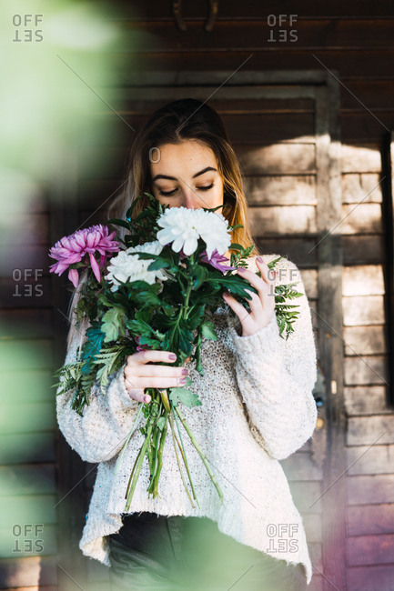 Beautiful content model in sweater smelling bunch of flowers standing on house porch