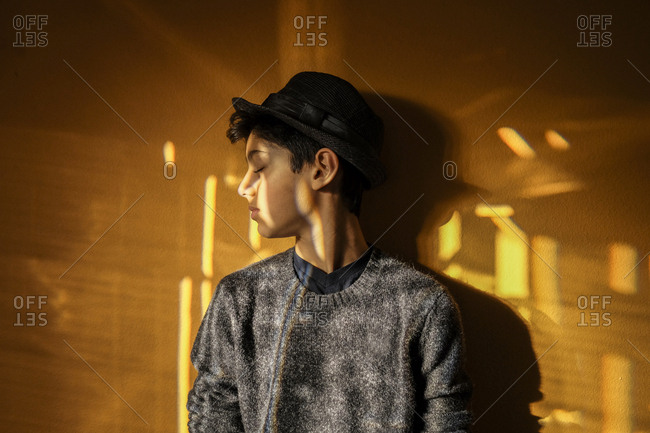 Boy wearing hat with eyes closed standing against yellow wall at home
