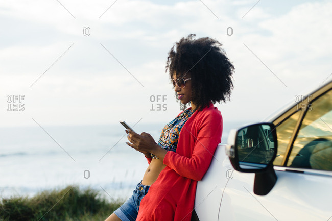 Fashionable afro hair woman on vacation texting on smartphone towards the sea. Stylish black model relaxing on a car trip to the coast.