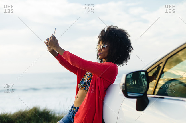 Fashionable afro hair woman on vacation taking selfie photo with smartphone towards the sea. Stylish black model on a car trip to the coast.