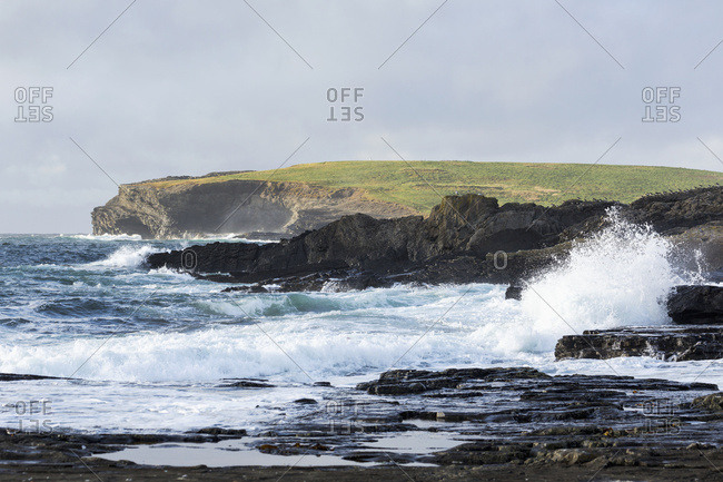 Waves Crashing Into Rocky Coast With Large Grassy Hill And Cliffs In Background; Kilkee, County Clare, Ireland