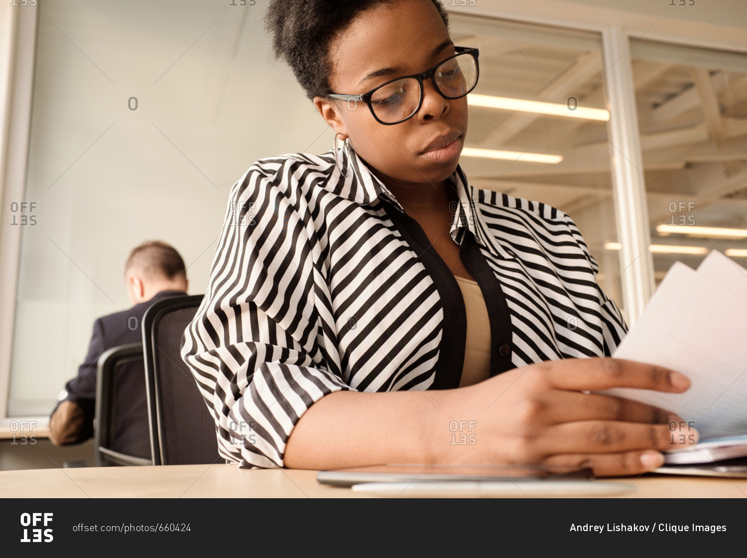 Low angle view of stylish black woman in striped blouse and glasses sorting documents while sitting at table in modern coworking space