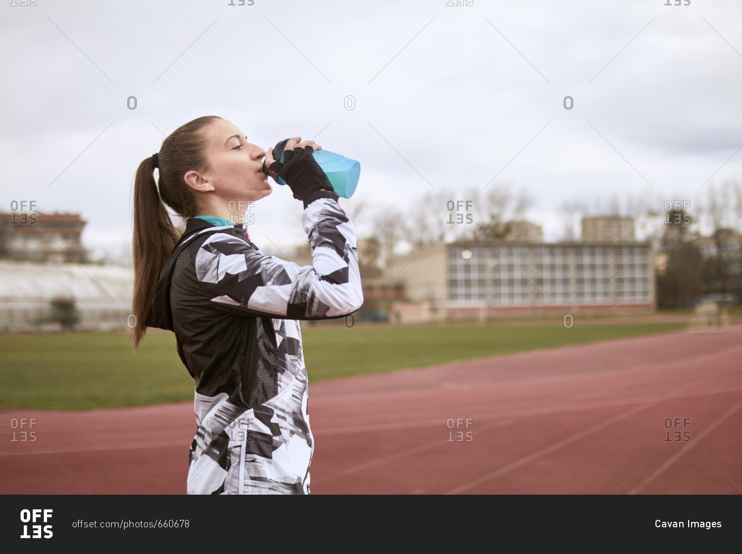 Side view of woman drinking water while standing against sky at sports track