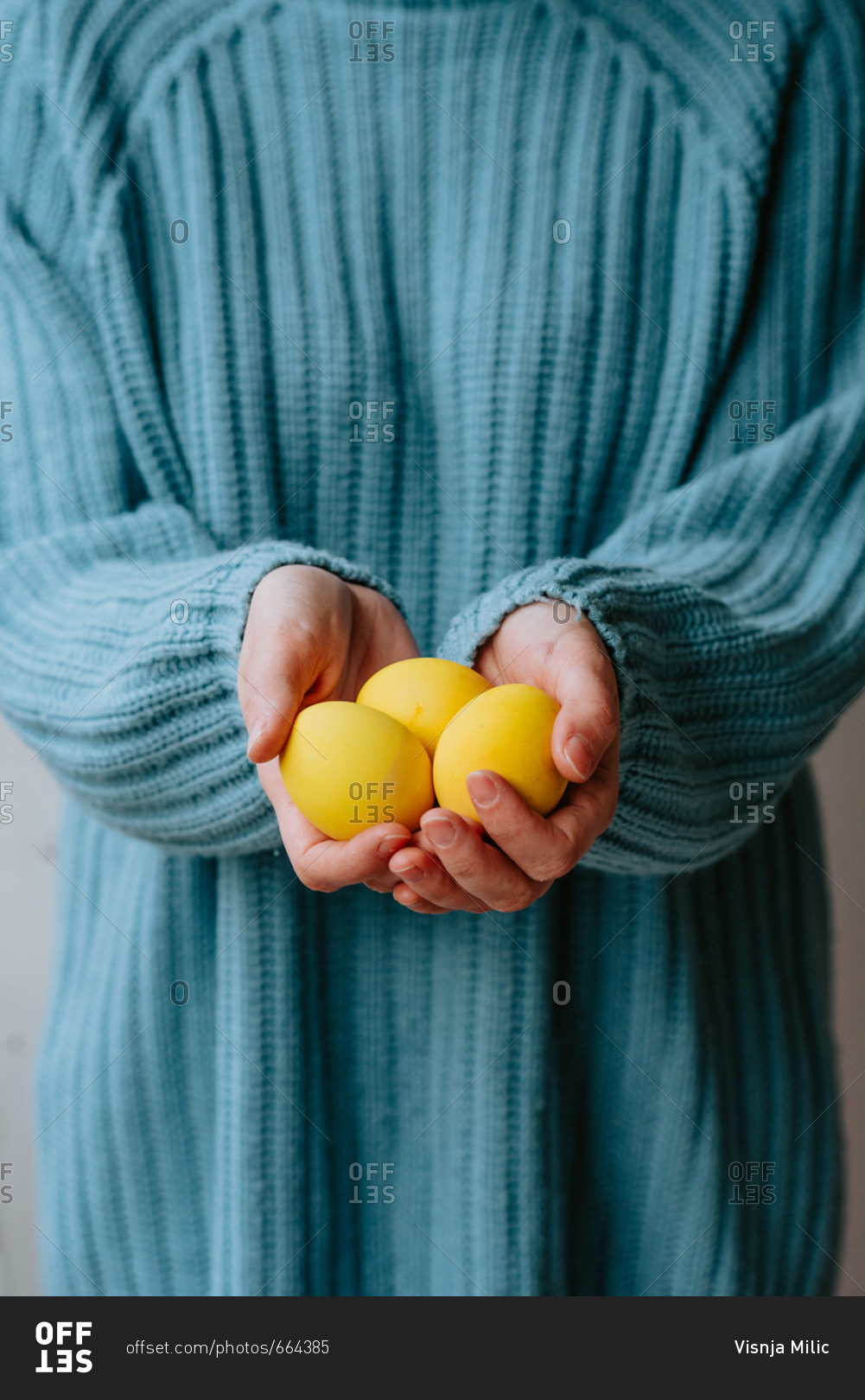 Person in pastel blue sweater holding three yellow Easter eggs