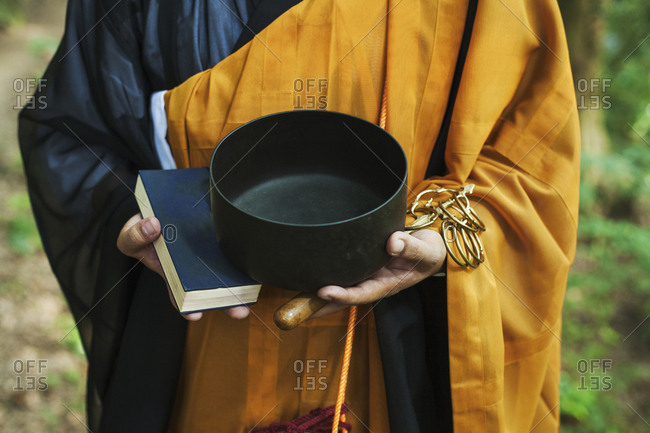 Close up of Buddhist monk wearing black and yellow robe, standing outdoors, holding prayer book and singing bowl