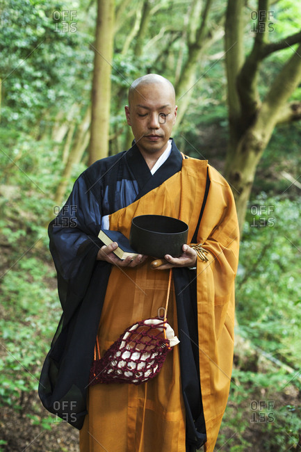 Buddhist monk with shaved head wearing black and yellow robe, standing outdoors, holding prayer book and singing bowl
