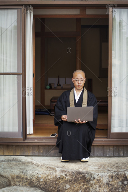 Buddhist monk with shaved head wearing black robe sitting outside temple, looking at laptop computer