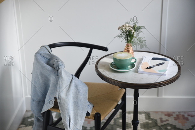 Denim jacket draped over back of chair at bistro table with coffee and notebook