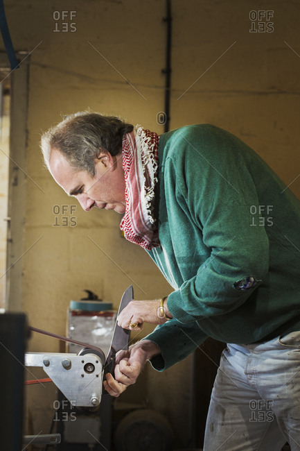 A craftsman in a knife makers workshop, holding a kitchen knife and shaping and finishing the wooden handle with a surface grinder a rotating wheel