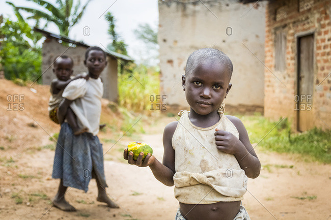 July 9, 2014: A Young Child Stands Holding A Piece Of Fruit With A Girl Holding A Young Child On Her Back In The Background; Uganda